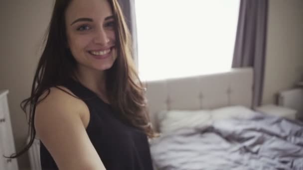 Slow motion footage of handsome cheerful girl enters a room and sit onto a bed,  selfie shot. — ストック動画