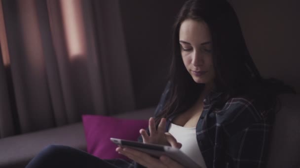 Handsome brunette girl sits on a couch indoors and uses tablet for business. — Αρχείο Βίντεο