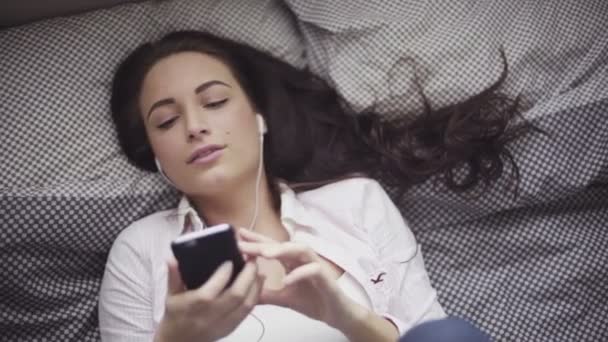 魅力的な白人の女の子はベッドの上で横になっていると自分のモバイル デバイスから、イヤホン接続で音楽を聴く. — ストック動画
