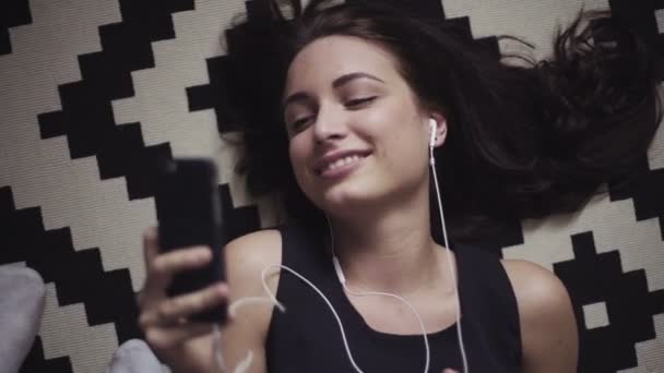 Retrato de una joven mujer tumbada en el suelo y hablando en su dispositivo móvil o escuchando la música con auriculares conectados . — Vídeos de Stock