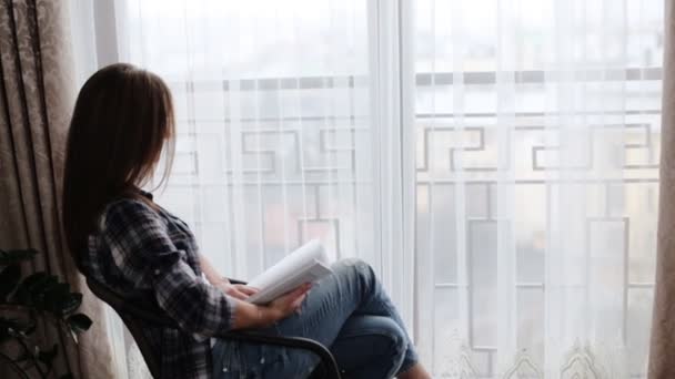 Nette Frau in blauen Jeans und grauem T-Shirt sitzt auf dem Tisch vor dem Fenster und liest in ihrem Wohnzimmer ein Buch. — Stockvideo