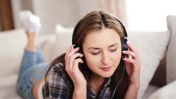 Junge schöne Frau auf dem Sofa liegend und Musik über Kopfhörer im Wohnzimmer hörend. — Stockvideo