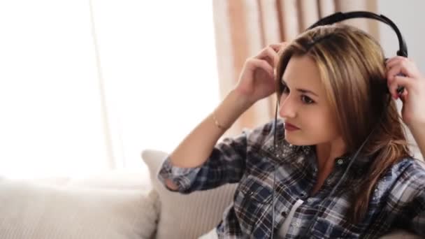 Joven hermosa mujer tumbada en el sofá y escuchando música en sus auriculares en la sala de estar . — Vídeos de Stock