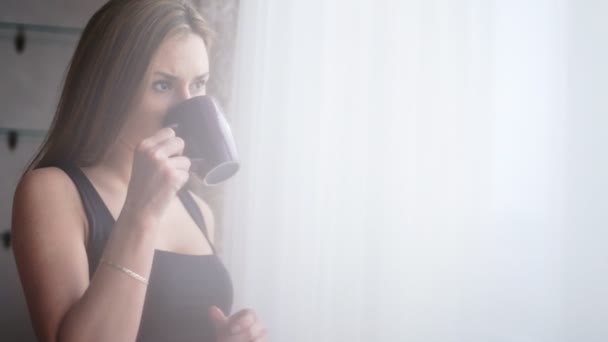 Atraktivní mladá dáma oddechuje a pití šálku čaje nebo kávy před okno doma. — Stock video