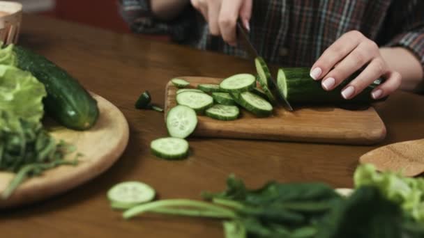 Jeune femme tranchant des concombres verts pour une salade avec un couteau, planche en bois . — Video