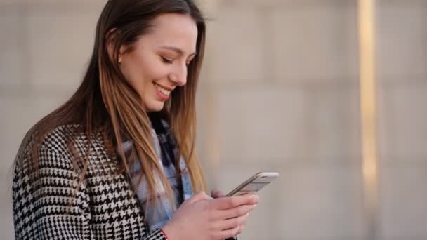 Mujer joven con abrigo de moda de colores blanco y negro usando su teléfono inteligente en la calle . — Vídeo de stock