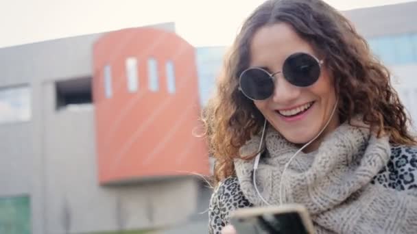 Adorable jeune femme souriante dans les écouteurs bavarder avec ses amis dans la rue . — Video