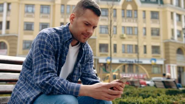 Profil eines glücklichen Kerls mit einem Smartphone, der auf einer Bank in einem Park sitzt — Stockvideo