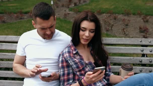 Couple sharing media in a smart phone sitting in a bench in a park with buildings in the background — Stock Video