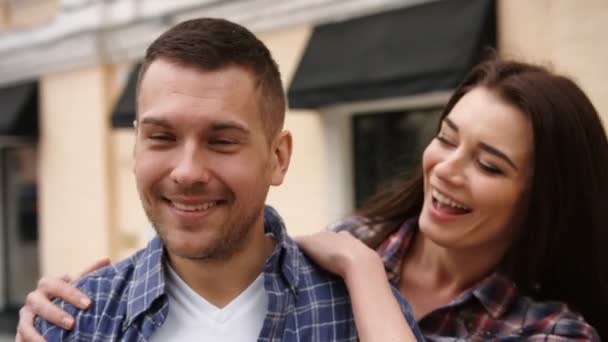 Giovane donna in camicia a scacchi che abbraccia il suo fidanzato sorridente da dietro — Video Stock