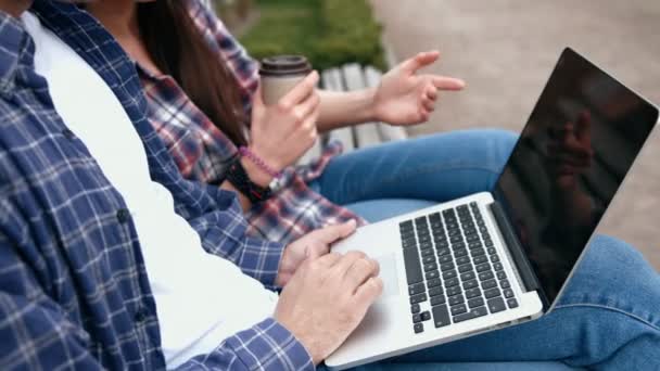 Jeunes citadins en chemises à carreaux et en jeans bleus utilisant un ordinateur portable et buvant du café de pour aller tasse sur banc — Video