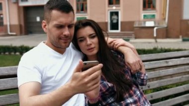 Young urban people man wearing white T-shirt and woman in checkered shirt with phones sitting on a bench