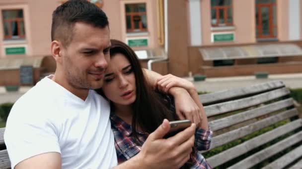 Young urban people man wearing white T-shirt and woman in checkered shirt with phones sitting on a bench — Wideo stockowe