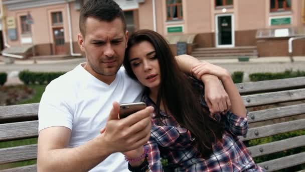 Joven urbano hombre vistiendo camiseta blanca y mujer con camisa a cuadros con teléfonos sentados en un banco — Vídeo de stock