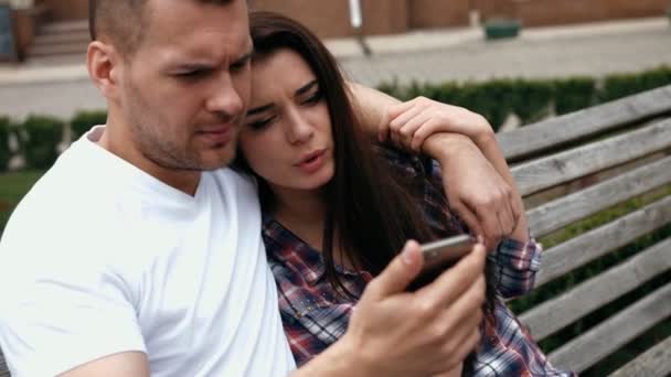 Giovani urbani uomo indossa maglietta bianca e donna in camicia a scacchi con i telefoni seduti su una panchina — Video Stock