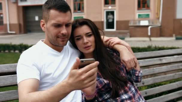 Young urban people man wearing white T-shirt and woman in checkered shirt with phones sitting on a bench — Stok video