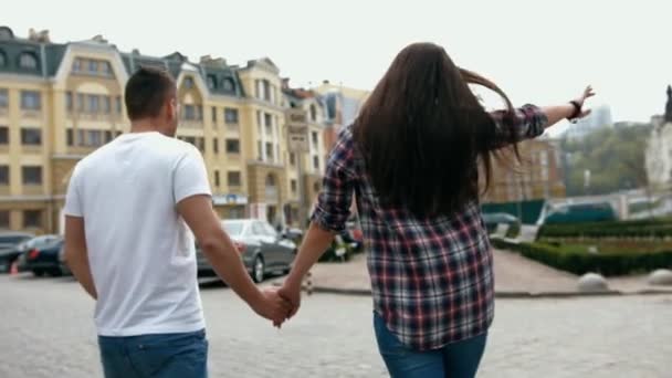 Jeune beau couple doux marchant la main dans la ville, portant un t-shirt blanc et chemise à carreaux, vue arrière — Video