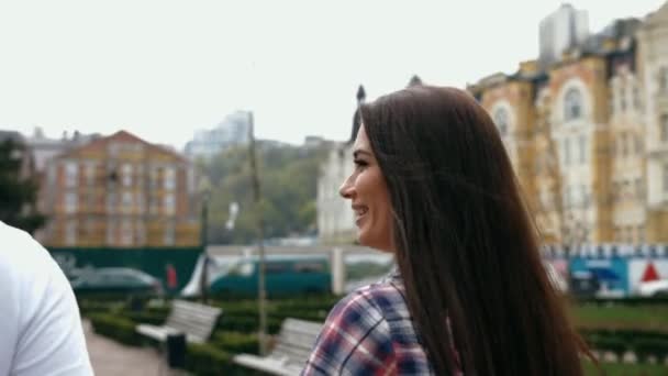 Hombre joven atractivo en camiseta blanca y mujer morena en una cita romántica, caminando cogidos de la mano, hablando y sonriendo feliz, mirándose el uno al otro, vista trasera — Vídeo de stock