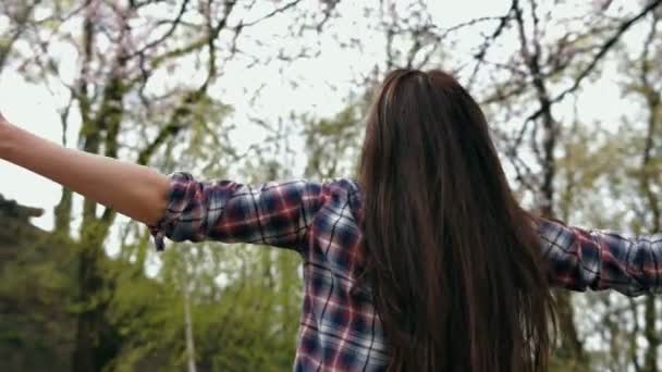 Feliz mujer morena sonriente con camisa a cuadros y vaqueros azules girando y riendo con los brazos extendidos al aire libre — Vídeos de Stock