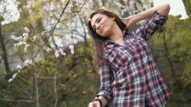 Çekici genç esmer kadın damalı gömlek elleri ile saç dokunmadan, parkta ağaçların önünde parlak güneşli bir günde açık havada ayakta