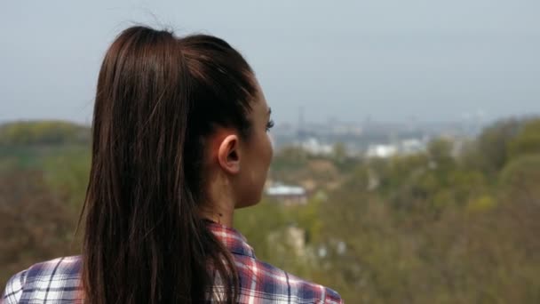 Primer plano de la joven mujer de cabello castaño con camisa a cuadros de pie de nuevo a la cámara mirando hacia fuera sobre exuberante paisaje urbano verde — Vídeo de stock