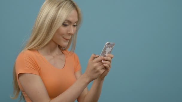 Sorprendido joven cabello rubio mujer feliz sosteniendo el teléfono móvil y mensajes de texto, mientras que de pie sobre fondo azul — Vídeos de Stock