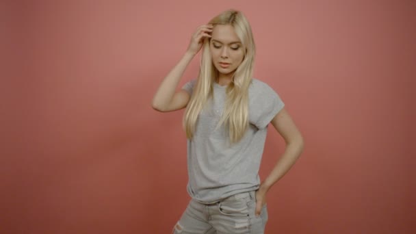 Retrato de una hermosa joven rubia con aspecto serio tocando el cabello y cruzando los brazos sobre fondo rosa — Vídeos de Stock