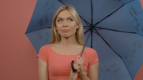 Verträumtes blondes Mädchen mit geheimnisvollem Lächeln, das wegschaut und einen blauen Regenschirm hält, isoliert auf rosa Hintergrund — Stockvideo