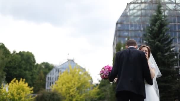 Casal romântico feliz, noiva sorridente e noivo com buquê rosa, dançando no jardim botânico, edifício de vidro no fundo — Vídeo de Stock