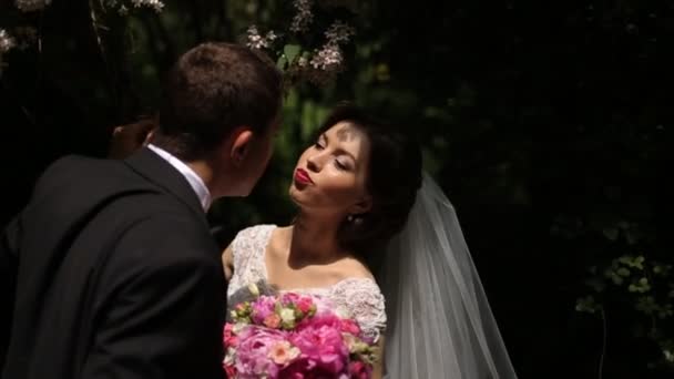 Novio guapo besando a la novia con ramo sobre un fondo de hojas de color verde oscuro, pareja tocándose las narices — Vídeos de Stock