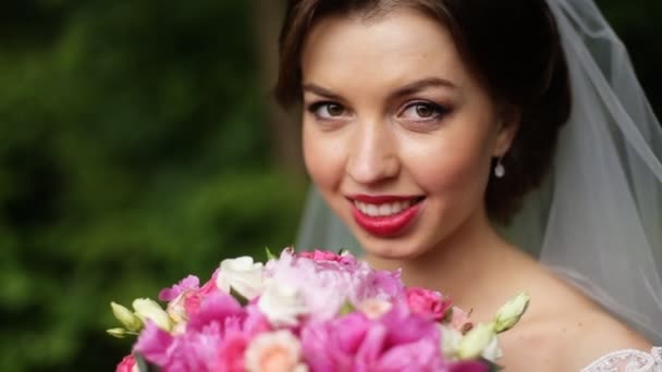 Portrait d'une mariée douce, sensuelle et très belle en robe blanche et voile, maquillage naturel, avec un bouquet de fleurs roses, gros plan — Video