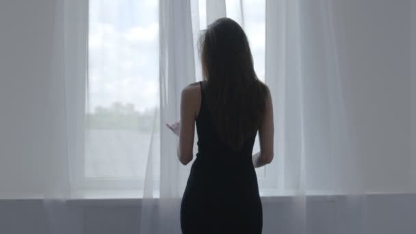 Mulher de vestido preto fica perto da janela e abre cortinas brancas, de volta para a câmera — Vídeo de Stock