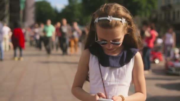 Blond kid girl playing with smartphone sitting on park lawn in city skyline background — Stock Video