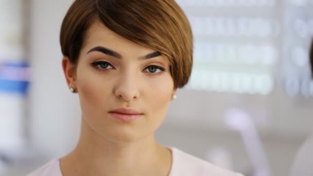 Fille romantique en chemise blanche élégante avec des cheveux bruns courts et un maquillage délicat souriant, regardant la caméra tout en posant contre le mur du studio blanc — Video