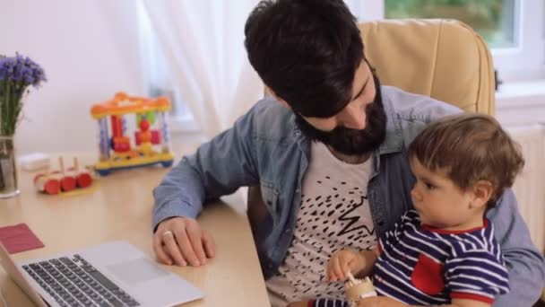 Tiempo en familia. Joven sonriente padre barbudo y su lindo hijo usando la computadora juntos en casa — Vídeo de stock