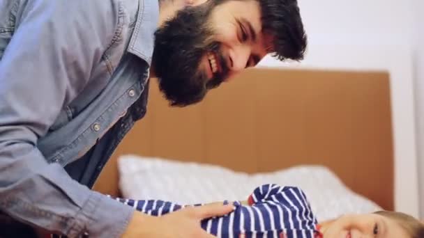 Menino sorridente deitado na cama brincando com seu pai barbudo — Vídeo de Stock