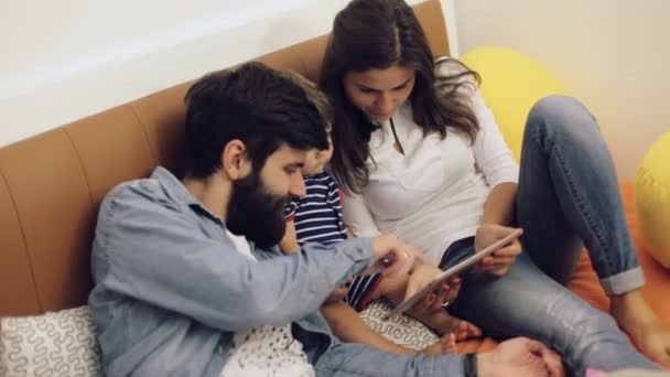 Pappa, mamma och deras unga son ha roligt genom att spela tillsammans på en tablett, de sitter på en soffa i vardagsrummet — Stockvideo