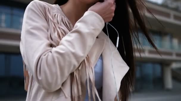 若い笑顔ブルネットの女性、ブルー ジーンズと白いジャケットを着てヘッドフォン、屋外に置くこと — ストック動画
