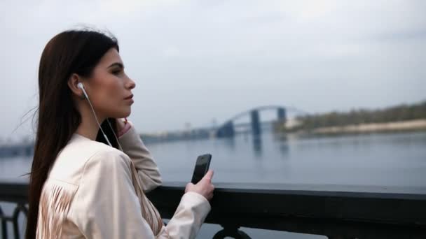 Mujer morena joven en chaqueta blanca escuchando la música con teléfono inteligente en el puente moderno. Ejercicio matutino en megaciudad — Vídeo de stock