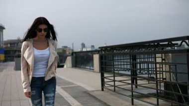 Genç ciddi esmer kadın beyaz ceket, kot ve modern köprüde yürürken siyah güneş gözlüğü.