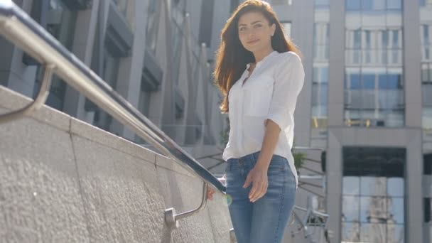 Крупним планом сексуальна брюнетка в білій сорочці і блакитні джинси, що спираються на металевий паркан на сучасній вулиці — стокове відео