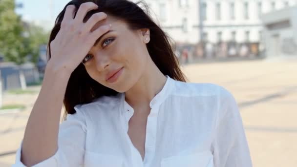 Atractiva joven morena con bonita cabeza temblorosa maquillaje, tocando el pelo por las manos, de pie al aire libre en un día soleado brillante frente a los edificios — Vídeos de Stock