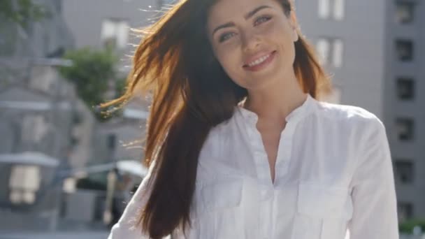 Hermosa joven morena posando en una ciudad soleada día de verano, en camisa blanca con el pelo largo soplado — Vídeos de Stock