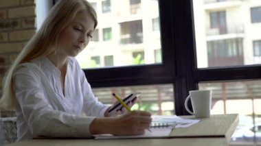 Kafede bir fincan kahve, not defteri ve tabletile ciddi genç bir kadın. Freelancer