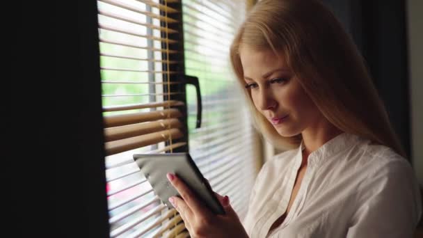 Primer plano de la encantadora mujer rubia sosteniendo la almohadilla táctil mientras está de pie cerca de la ventana en el interior moderno — Vídeos de Stock