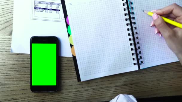 Foto aérea de una mujer escribiendo en un bloc de notas. Hay un teléfono inteligente con una pantalla verde — Vídeo de stock