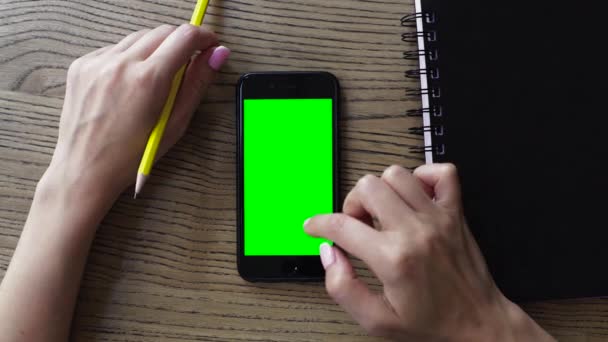 Main féminine utilisant un téléphone portable intelligent avec un écran vert sur une table en bois, vue aérienne — Video