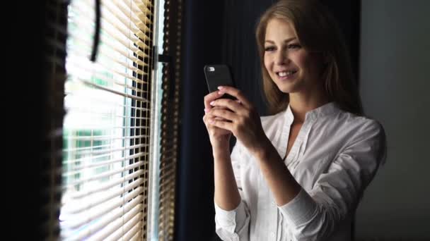 Mujer rubia joven con camisa blanca usando el teléfono celular mientras está de pie junto a una ventana — Vídeos de Stock