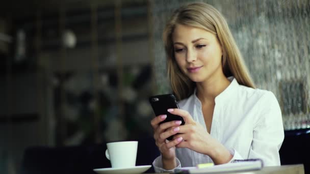 Porträt einer niedlichen blonden Frau, die in einem modernen Café etwas auf ihrem Smartphone liest — Stockvideo