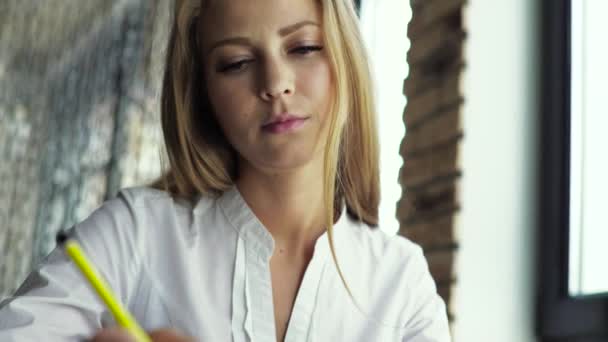 Блондинка сидит за столом, пишет в блокноте и пьет кофе в приятном светлом интерьере — стоковое видео